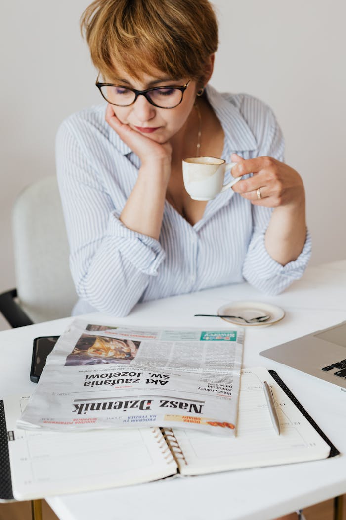 A thoughtful woman reads a newspaper while enjoying coffee at an indoor workspace.