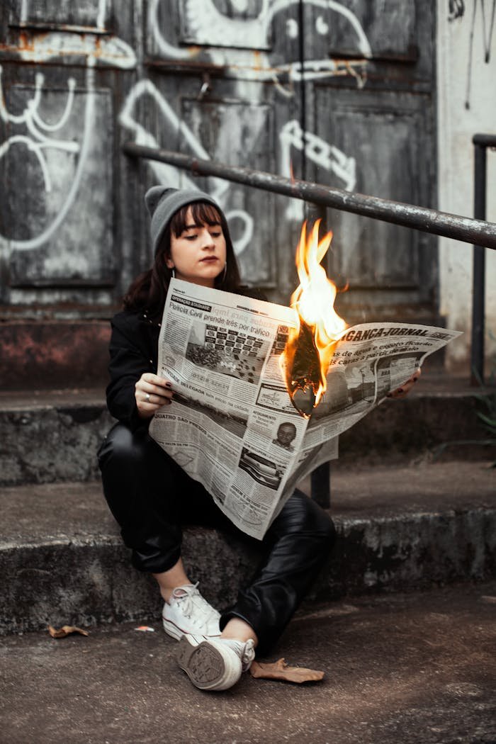 team-03 Woman sits outdoors reading a newspaper on fire, creating a dramatic urban scene.