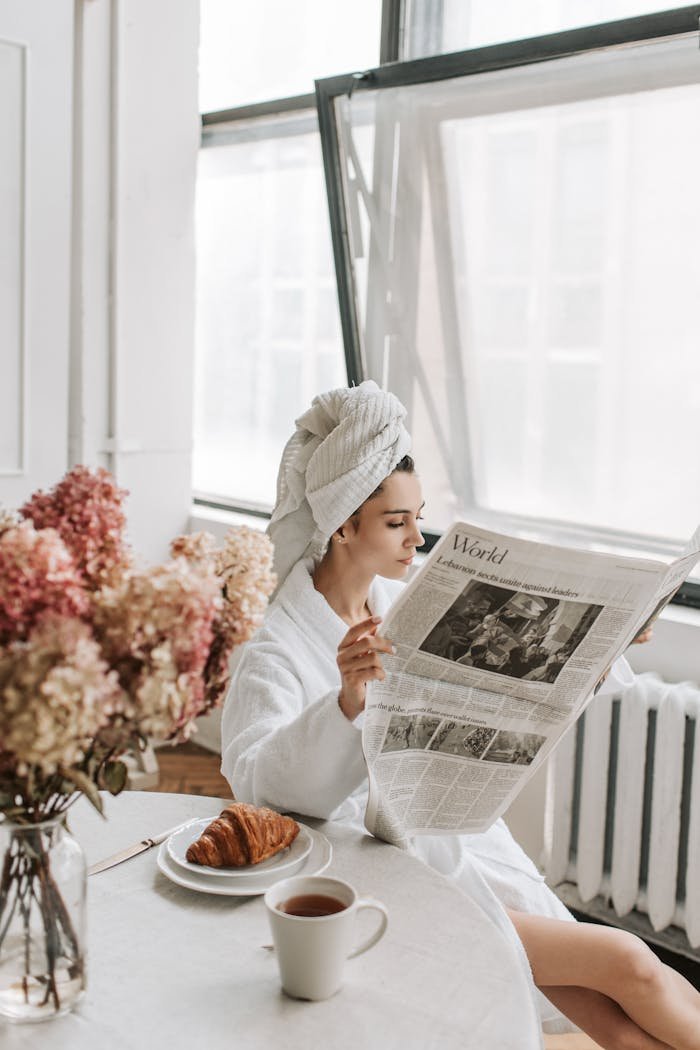 team-04 A woman in a bathrobe reads a newspaper while enjoying breakfast indoors with coffee and croissants.