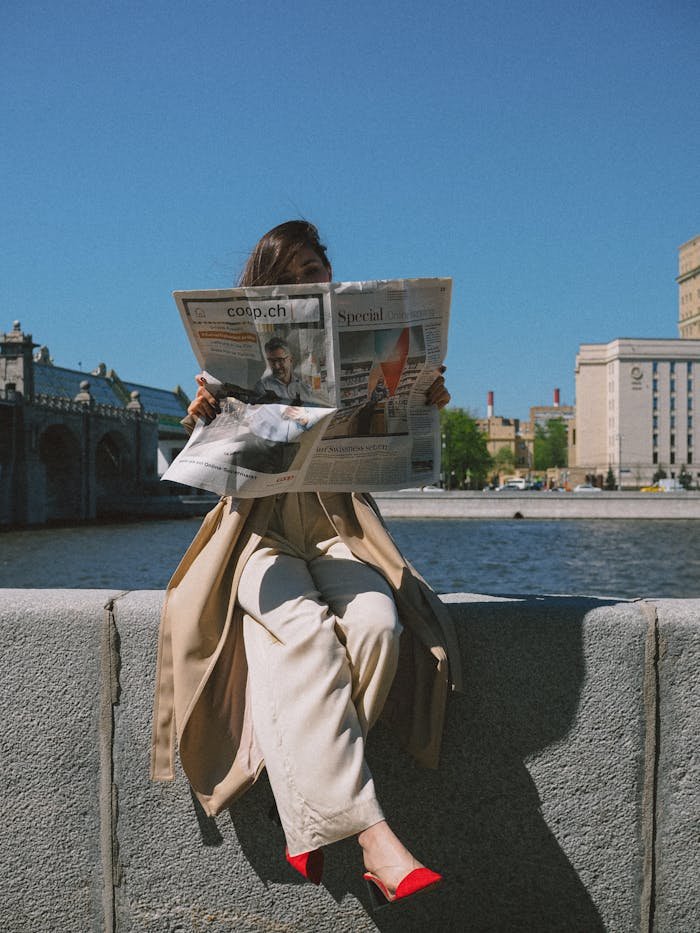 team-02 Woman sitting by river holding newspaper, city backdrop, sunny day.